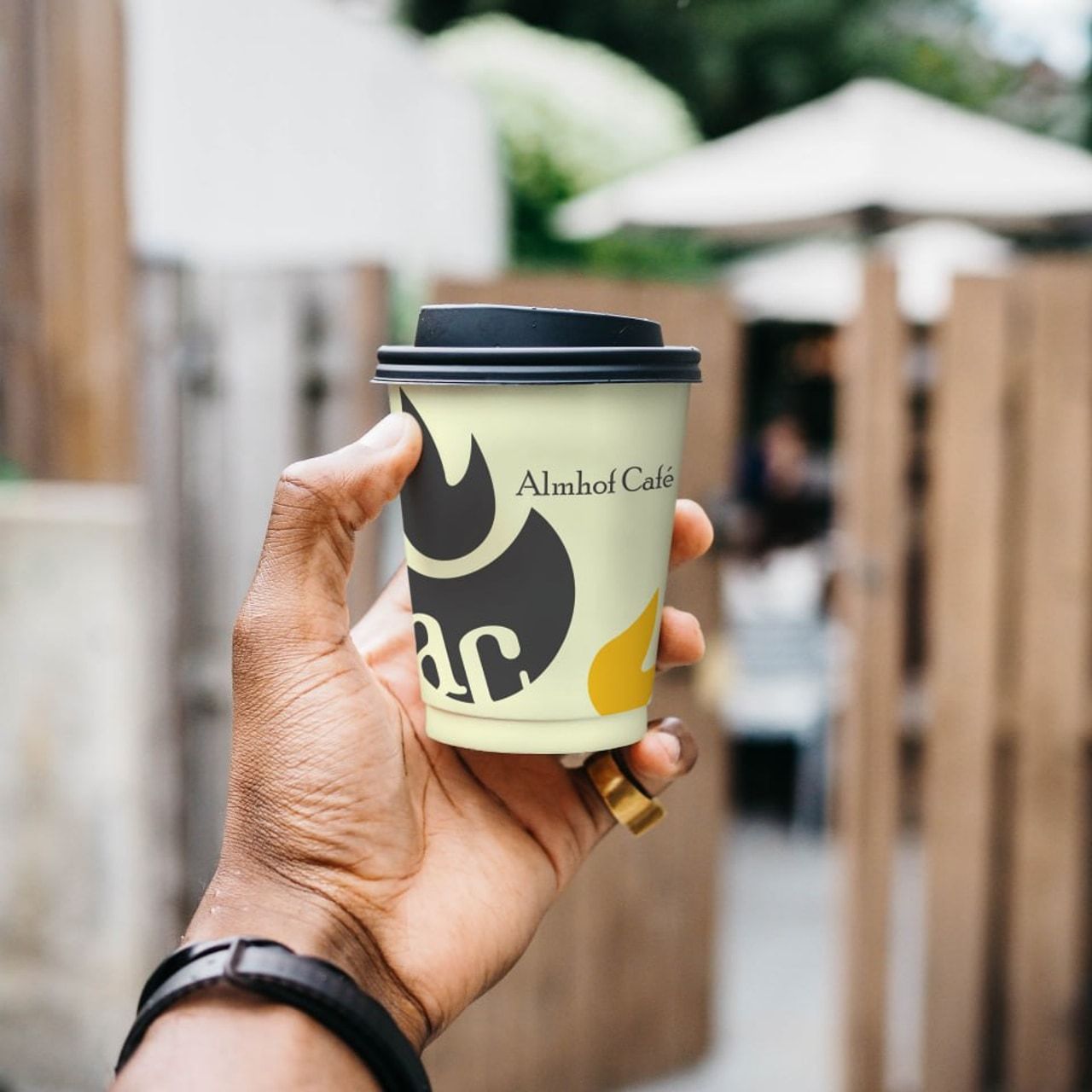 Стаканчик с логотипом Almhof Café в руке на фоне размытой уличной застройки